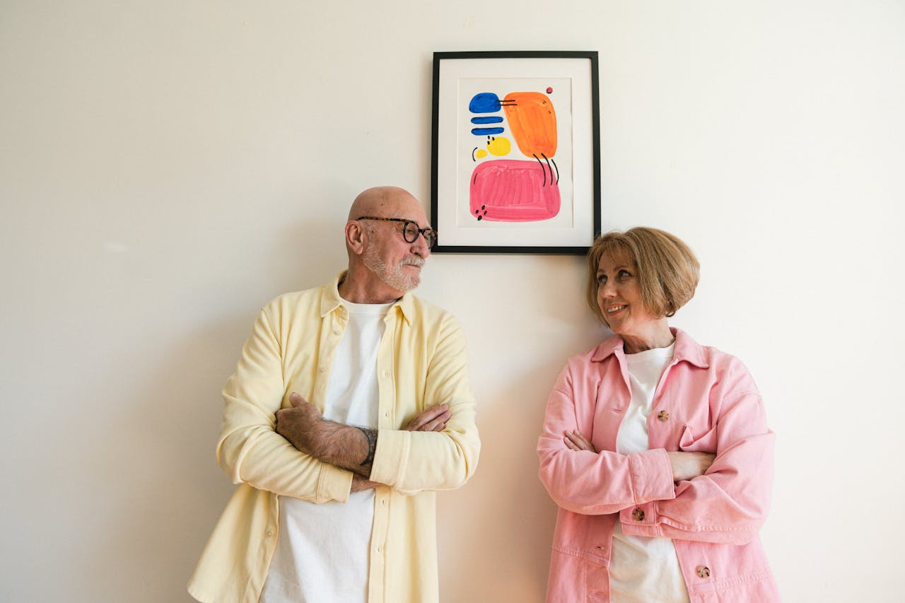 Elderly couple stands smiling at each other under vibrant artwork on a plain wall indoors.